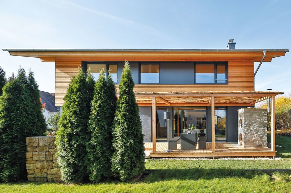 Modernes zweistöckiges Haus mit Holzverkleidung, großen Fenstern und einer Veranda, umgeben von grünen Bäumen und einer Steinmauer.