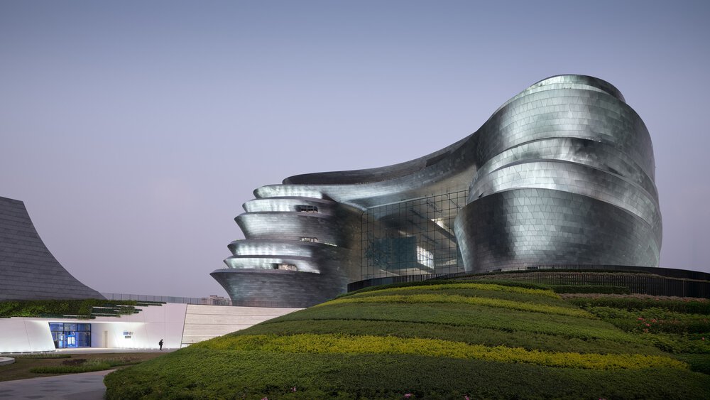 A large, futuristic, curved silver building with a large glass front on a green hill at dusk. A person is standing in the entrance area.