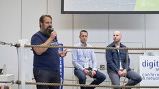 A man is speaking into a microphone in a boxing ring, behind him two other men are sitting on chairs next to a desk labeled “conference & networking”.