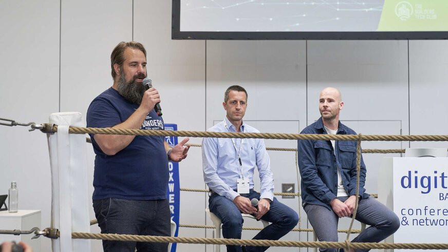 A man is speaking into a microphone in a boxing ring, behind him two other men are sitting on chairs next to a desk labeled “conference & networking”.