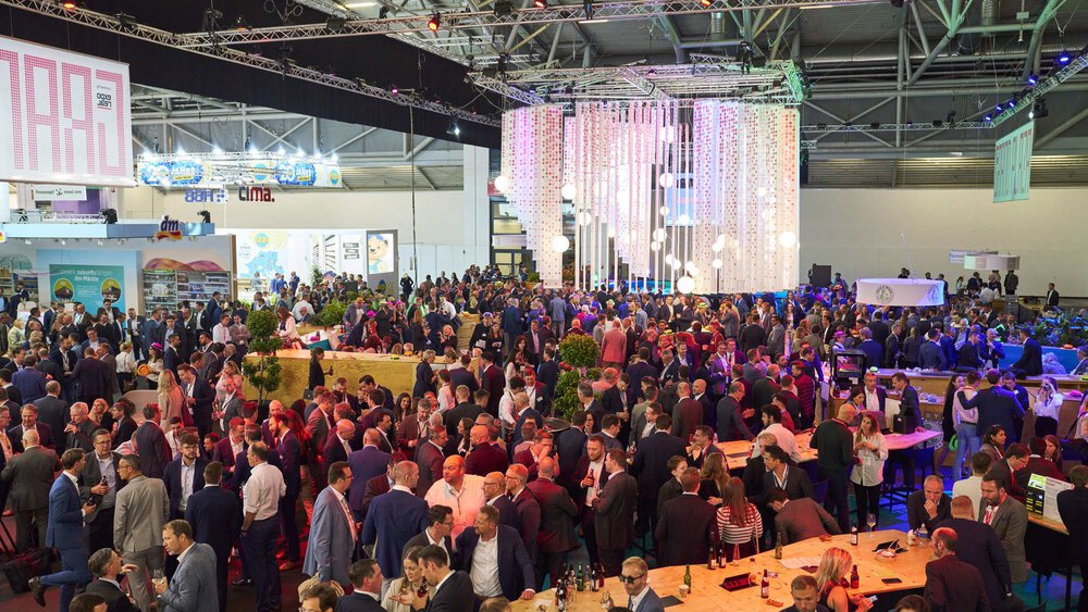 A large crowd gathers in an exhibition hall, in an area with large tables and counters. People are drinking and chatting.