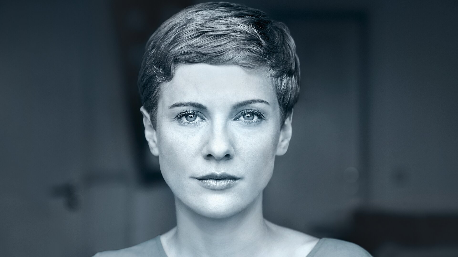 Eine Frau mit kurzen Haaren und ernstem Gesichtsausdruck blickt direkt in die Kamera in einer gedämpften, blau getönten Umgebung.