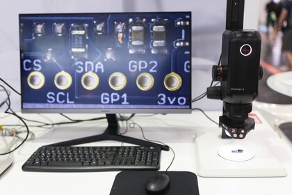 Close-up of an electronic circuit board with labeled components, next to a microscope and a keyboard on a desk.