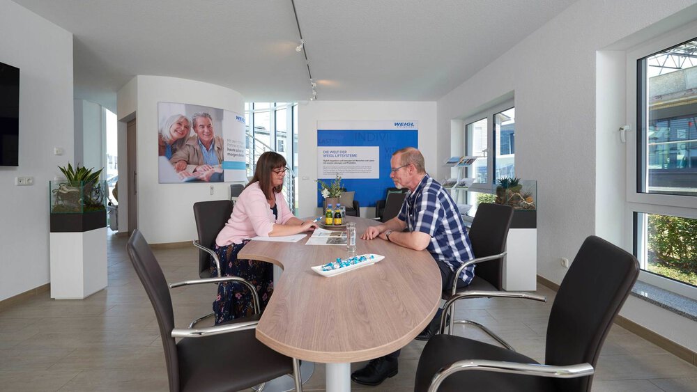 Eine Frau und ein Mann sitzen sich an einem Tisch in einem modern eingerichteten Büro mit großen Fenstern gegenüber.