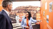 A man in a suit and a dark-haired woman stand in front of a background image of a container and an astronaut in a red rocky desert. The woman points to a presentation wall in orange and talks to the man.
