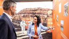 Ein Mann im Anzug und eine dunkelhaarige Frau stehen vor einem Hintergrundbild mit einem Container und einem Astronauten in einer roten Felswüste. Die Frau deutet auf eine Präsentationswand in Orange und unterhält sich mit dem Mann.