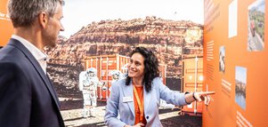Ein Mann im Anzug und eine dunkelhaarige Frau stehen vor einem Hintergrundbild mit einem Container und einem Astronauten in einer roten Felswüste. Die Frau deutet auf eine Präsentationswand in Orange und unterhält sich mit dem Mann.