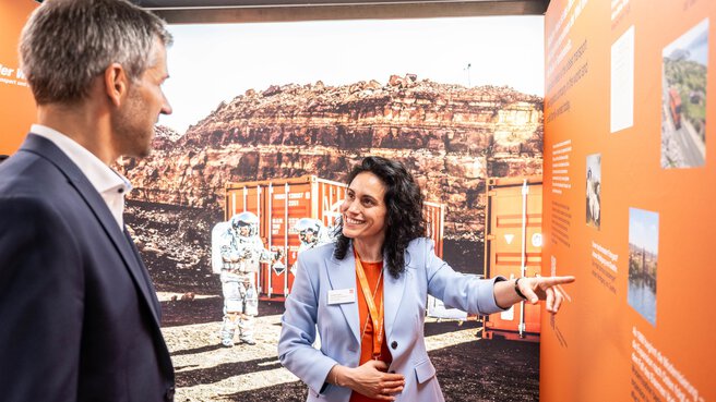A man in a suit and a dark-haired woman stand in front of a background image of a container and an astronaut in a red rocky desert. The woman points to a presentation wall in orange and talks to the man.