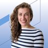Eine lächelnde Frau mit langen gelockten braunen Haaren trägt einen blau-weiß gestreiften Pullover und Ohrringe.