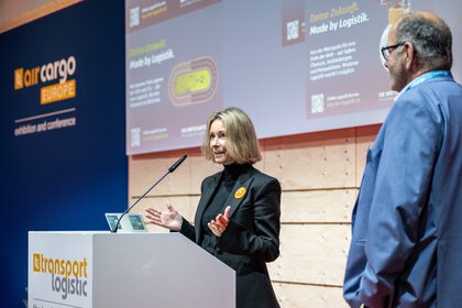 A woman speaks on a podium while a man stands next to her at a transportation logistics conference.