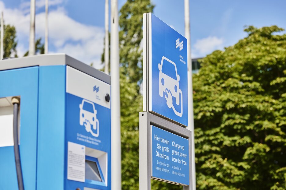 A blue charging station for electric cars with signage, outdoors with trees and a blue sky in the background.