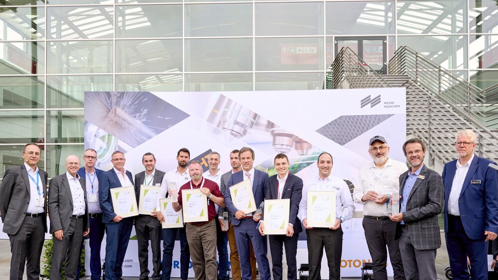 Eine Gruppe von Menschen posiert mit Auszeichnungen vor einer Messehalle und einem Plakat der World of Photonics.