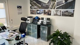 Modernes Büro mit Glastisch, Bürostuhl, Drucker, Schränken, Topfpflanze und Fotos an der Wand.