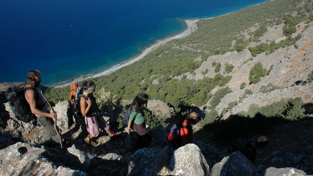 Five hikers descend a rocky mountain path, looking down at the blue sea and the wooded coastline below them.