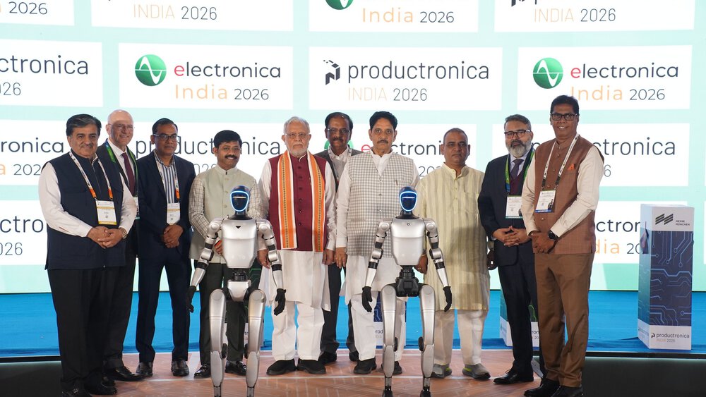 A group of people and two humanoid robots are posing on stage at productronica India 2026.