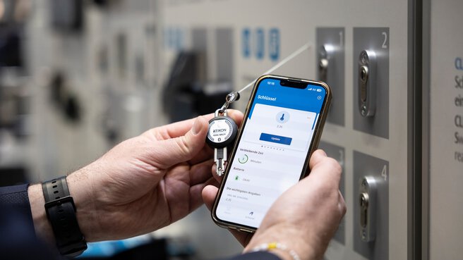 Zwei Hände halten in der linken Hand einen Schlüssel und in der Rechten Hand ein Smartphone. Auf dem Smartphone ist eine Sicherheitssoftware zu sehen, die den Schlüssel erkennt.