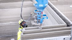 A construction worker wearing a helmet and mask operates a large blue 3D concrete printer.