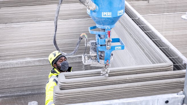 Ein Bauarbeiter mit Helm und Maske bedient einen großen blauen 3D-Betondrucker.