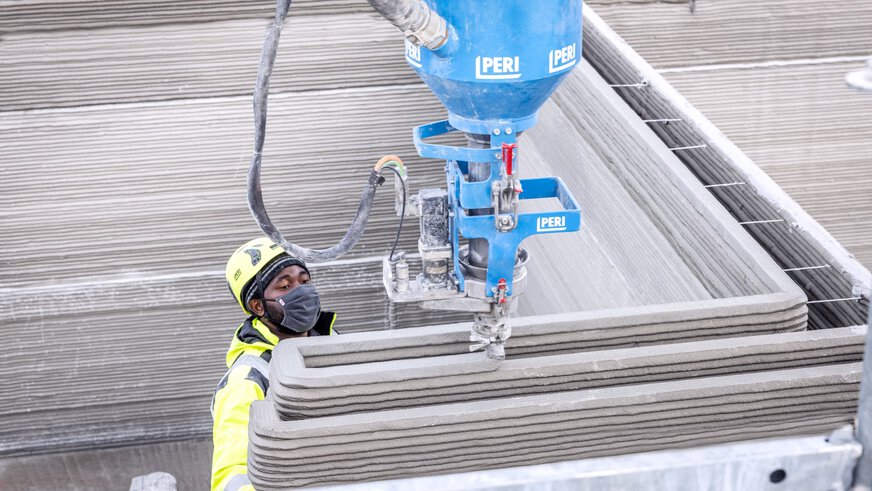A construction worker wearing a helmet and mask operates a large blue 3D concrete printer.