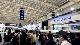 A crowded exhibition hall where people are walking among brightly lit booths and exhibition areas displaying various company signs and products.