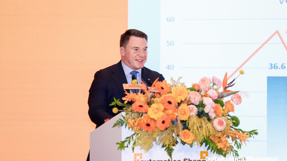 Man giving a presentation at a podium at automatica Shanghai with a slide and floral arrangement in front.