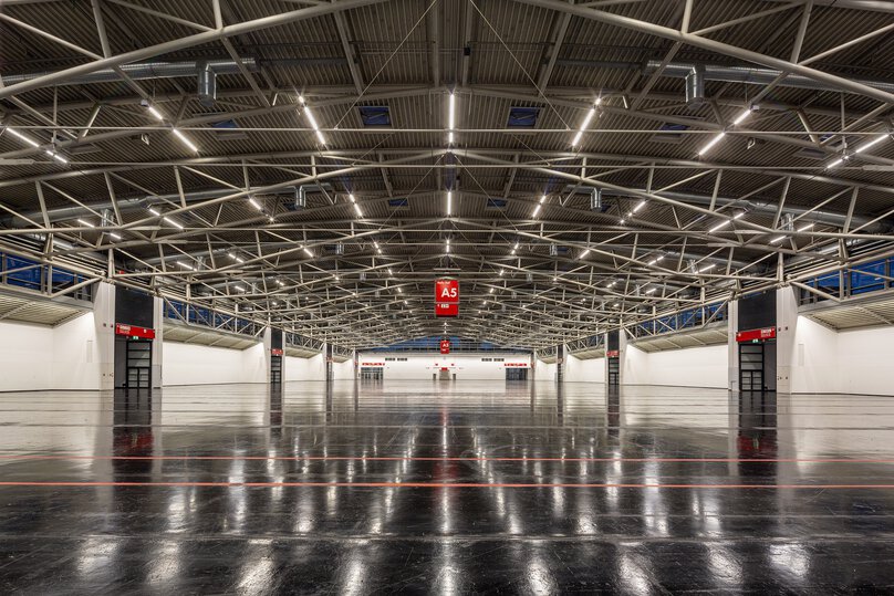 A spacious, empty industrial hall with exposed metal beams, a polished dark floor and red A5 signs hanging above.