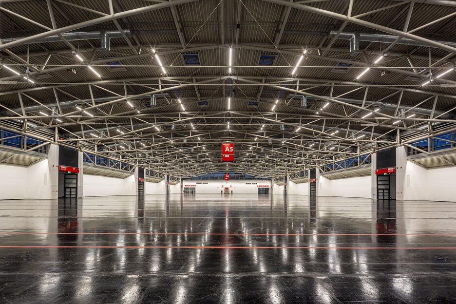 A spacious, empty industrial hall with exposed metal beams, a polished dark floor and red A5 signs hanging above.