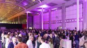 A large crowd gathers with drinks at bar tables in a large hall with purple lighting.