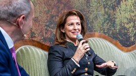 Eine Frau, die in ein Mikrofon spricht, während sie neben einem Mann auf einem Sofa sitzt.