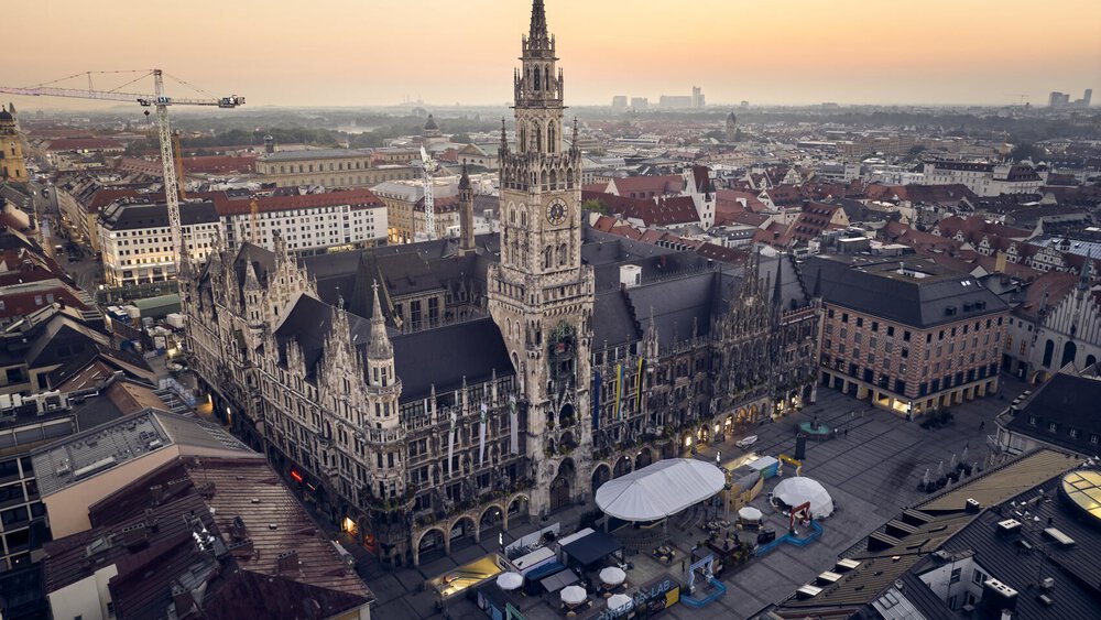 Luftaufnahme des Neuen Rathauses in München bei Sonnenuntergang, umgeben von städtischen Gebäuden und einem offenen Platz.