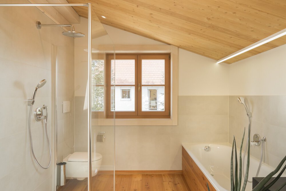 Modernes Badezimmer mit Glasdusche, WC, Badewanne und schräger Holzdecke; Fenster mit Blick ins Freie.