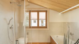 Modernes Badezimmer mit Glasdusche, WC, Badewanne und schräger Holzdecke; Fenster mit Blick ins Freie.