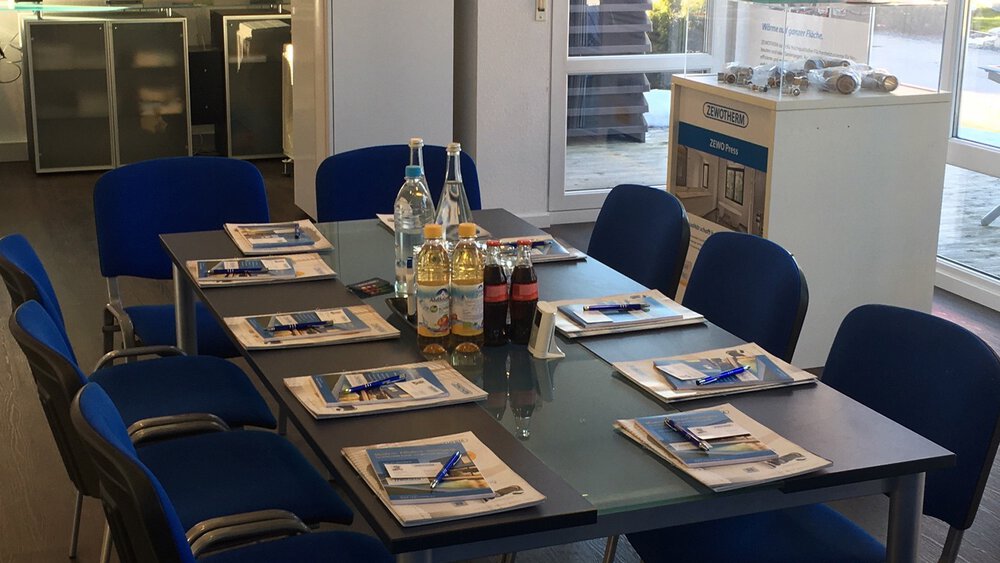 Besprechungsraum mit Tisch, blauen Stühlen, Notizblöcken, Stiften und Getränken. Im Hintergrund Regale und Fenster.