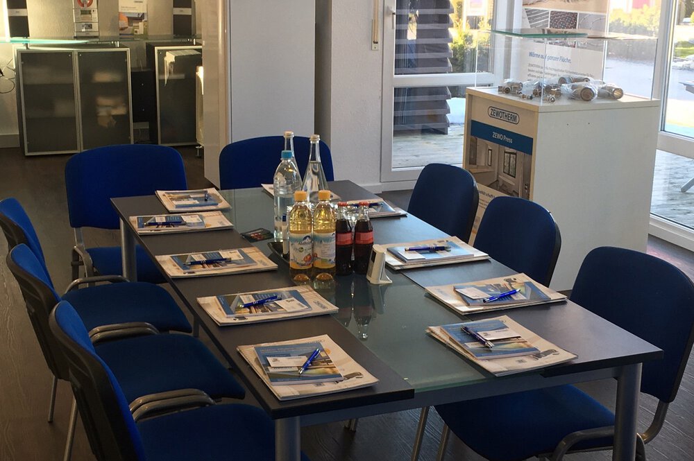 Besprechungsraum mit Tisch, blauen Stühlen, Notizblöcken, Stiften und Getränken. Im Hintergrund Regale und Fenster.