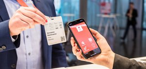 Eine Person scannt mit einem Smartphone mit roter App-Oberfläche einen QR-Code auf einem Besucherausweis.
