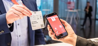 A person scans a QR code on a visitor badge using a smartphone with a red app interface.