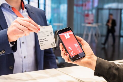 A person scans a QR code on a visitor badge using a smartphone with a red app interface.