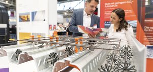 Nahaufnahme eines Modells aus Metallschienen, Streben und Ringabstandhaltern an einem Messestand der BAU. Dahinter unterhält sich ein Aussteller mit einer Besucherin.