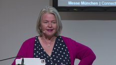 Eine grauhaarige Frau spricht in einem Headset-Mikrofon vor einem Bildschirm.