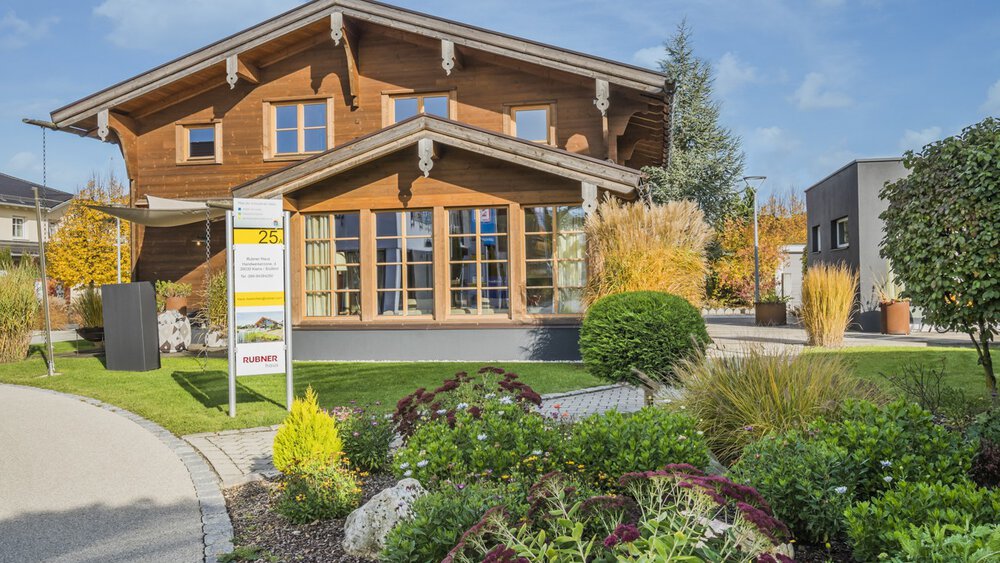 Holzhaus mit großen Fenstern, umgeben von einem üppigen Garten und einem angelegten Gehweg unter einem klaren blauen Himmel.