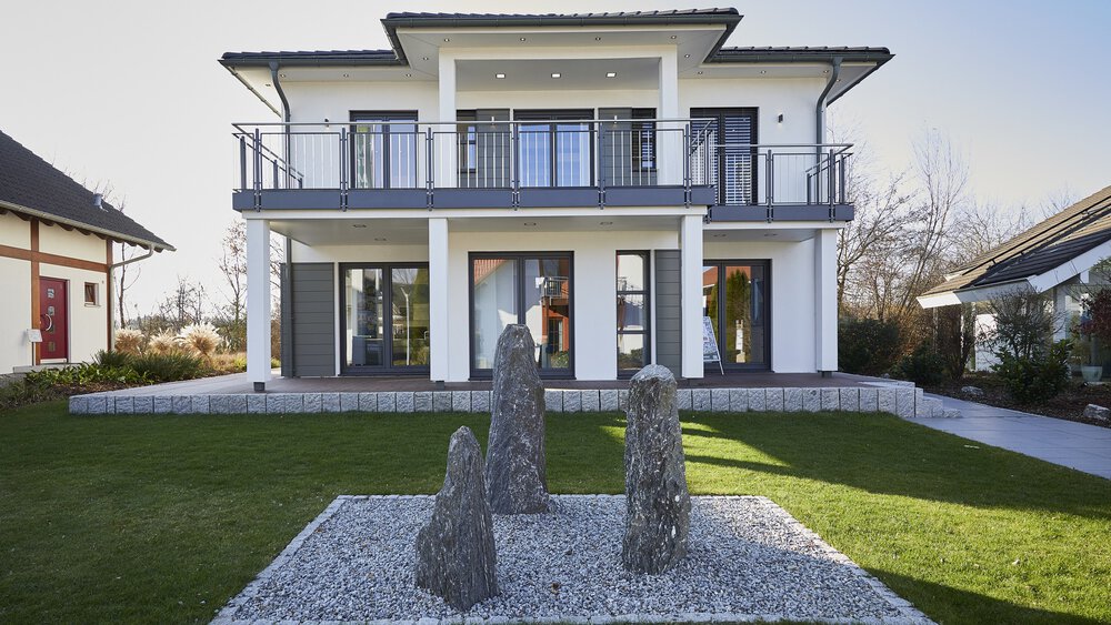 Modernes zweistöckiges Haus mit großen Fenstern, Vorgarten und Hecken unter einem klaren blauen Himmel.