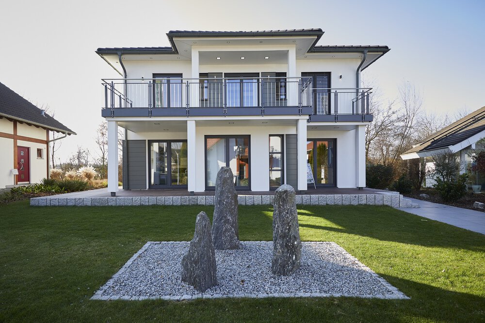 Modernes zweistöckiges Haus mit großen Fenstern, Vorgarten und Hecken unter einem klaren blauen Himmel.