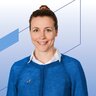 Eine lächelnde Frau mit hochgesteckten rötlichen Haaren trägt eine weiße Bluse und einen blauen Cardigan darüber.
