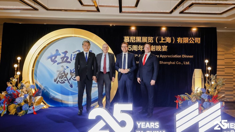 Four men in suits stand in front of a press wall with decorations on the sides and a display with the words “25 YEARS IN CHINA” in front of them.