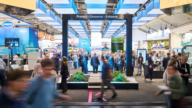 Ein moderner, offen gestalteter Messestand unter blau-weißen Leuchtelementen mit dem Slogan „Protect - Conserve - Enhance“ und zahlreichen Besuchern.