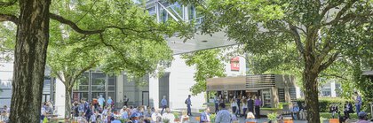 On a sunny day, people eat and chat at tables under trees on a green area between the exhibition halls.