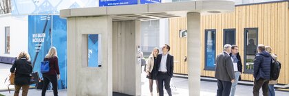 Menschen stehen um einen modernen Betonunterstand mit Bannern, der vor einer Messehalle ausgestellt ist.
