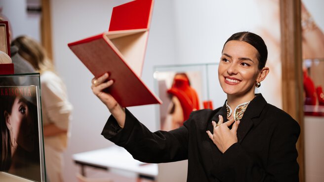 A woman in a black jacket wears a piece of jewelry around her neck and looks at herself smiling in a large red mirror that she holds in her hand.