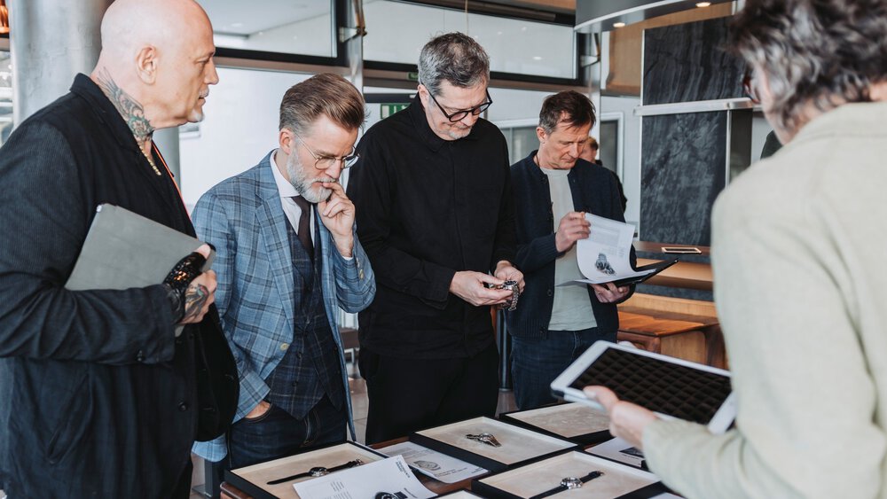 Several people are standing together around a table with many black wooden boxes in which various wristwatches are displayed.