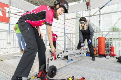 Two women in work clothes lift a folded scaffolding system by two straps.